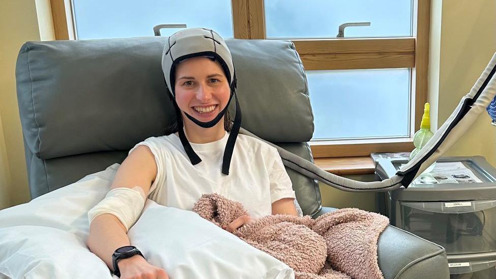 Lucy Fitton sitting in hospital while having a chemotherapy treatment. She is sitting in a large grey chair with one arm resting on a pillow and has a pink blanket over her legs. She is wearing a light grey chemotherapy cold cap on her head. 