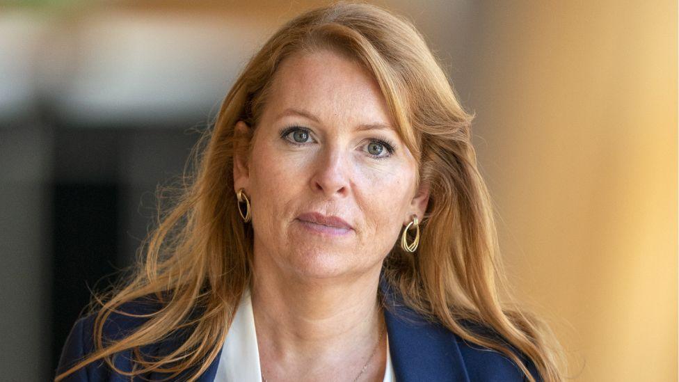 Ash Regan, a red-haired woman wearing a blue jacket and white shirt, looks at the camera in a medium close-up. She is walking in a Scottish Parliament corridor, which is blurred in the background. 