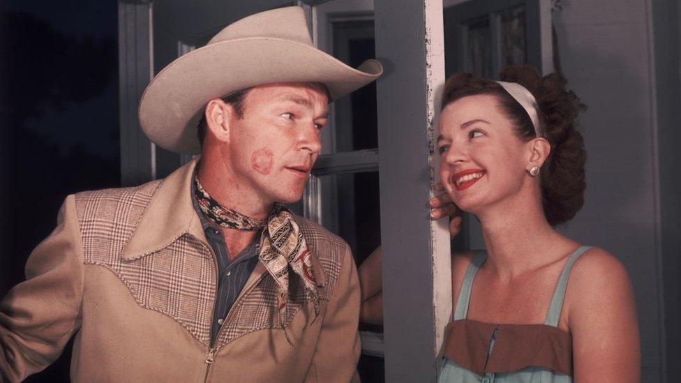 Dale Evans (1912 - 2001) smiles after leaving a red lipstick mark on the cheek of her husband, American singer and actor Roy Rogers (1911 - 1998), upon his arrival through the front door