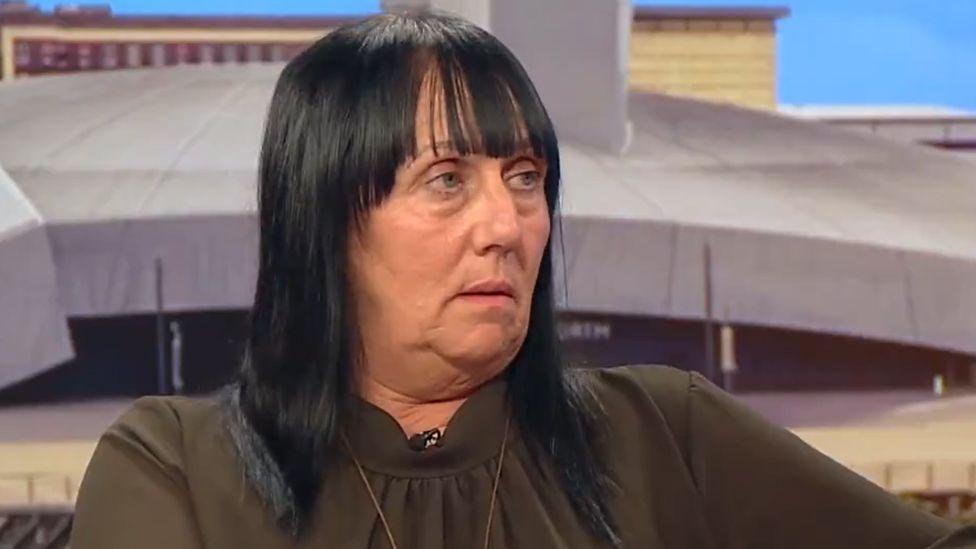 Debbie Duncan sits on a sofa at BBC Breakfast. The head and shoulders image shows her looking to the right at the presenter. She is wearing a dark brown top and she has long, shiny, straight black hair with a fringe.