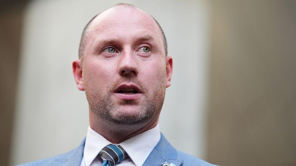 A bald man in a blue suit, white shirt and blue stripy tie looks to his left in a close-up shot 