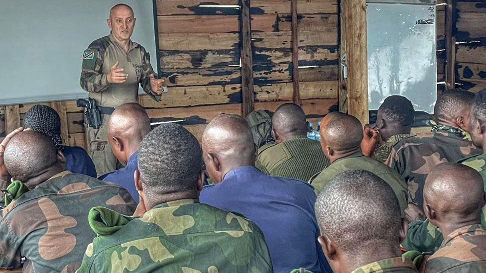 Horațiu Potra in military fatigues and a holstered gun stands in front of a class full of Congolese soldiers