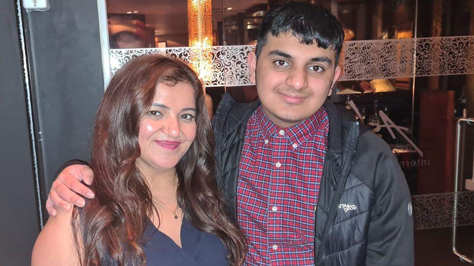 Pooja Kanda, wearing a blue dress, poses with her teenage son Ronan.  He has his arm around her shoulder (he is a bit taller) and wears a check shirt and black coat. Both smile. 