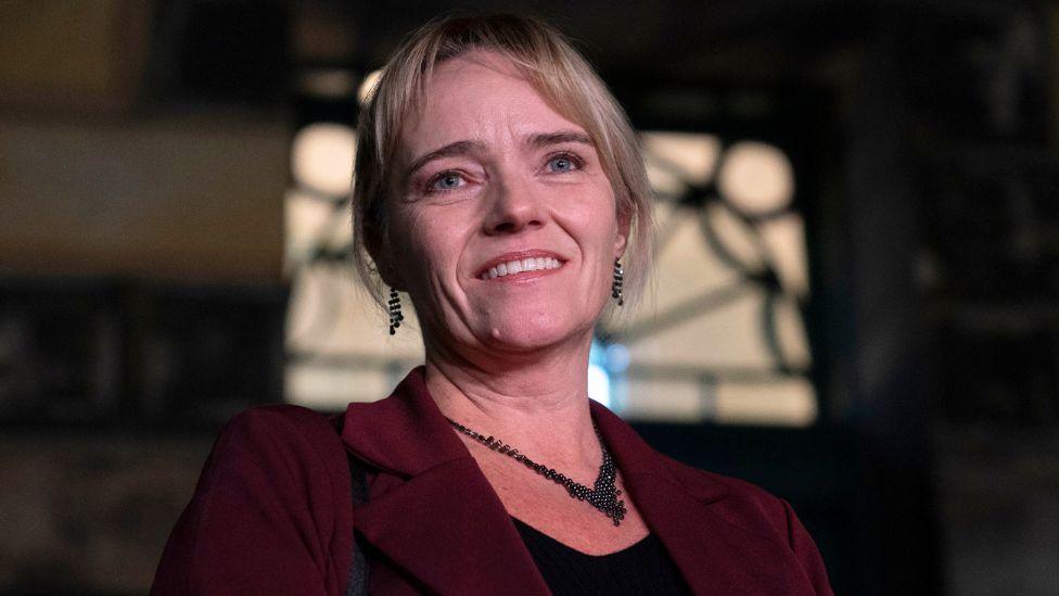 A portrait of a woman, shown from the shoulders up, with a dark background and soft lighting. She has medium-length blonde hair that is neatly styled.