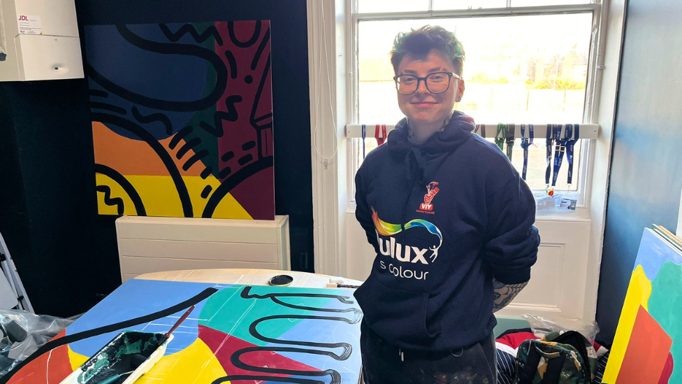 A woman with short hair and glasses, wearing a Dulux jumper, stands next to a colourful mural.