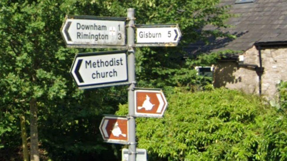 The original sign shows two signs pointing to the left saying "Downham 1, Rimington 3" on one and "Methodist Church" on one below it. There is a brown sign with a white witch icon underneath and also one to the right under another sign pointing to the right saying "Gisburn 5".