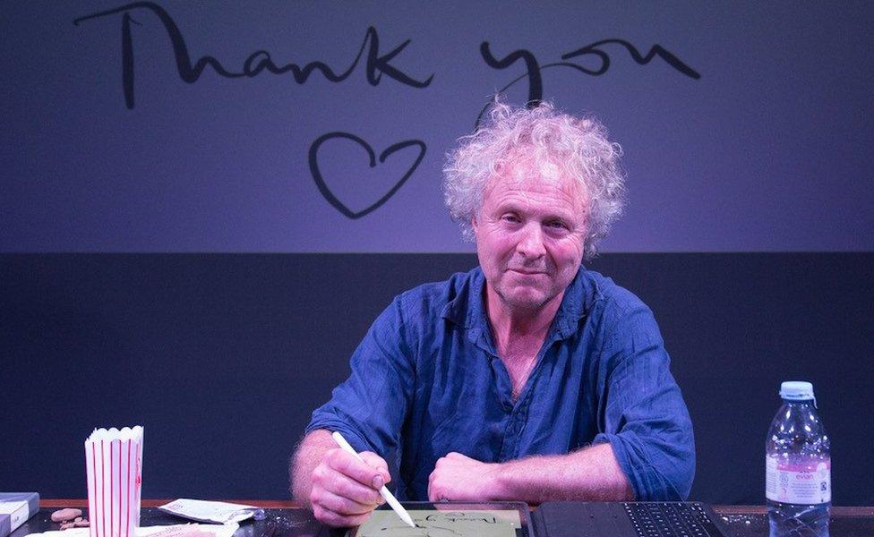 Charlie Mackesy sitting at a desk on stage and drawing