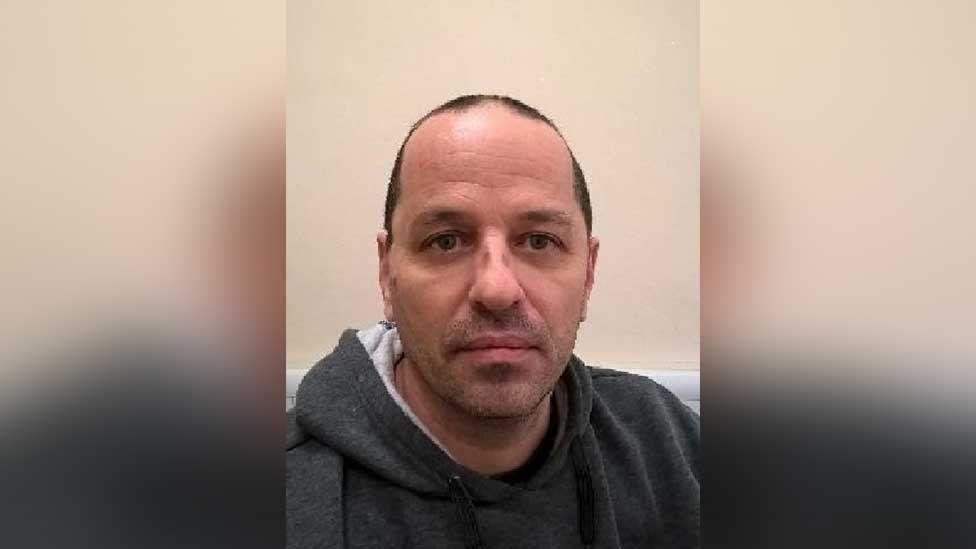 A police custody photo of Neil Maxwell, a middle-aged man with short black hair who is wearing a prison-issue grey hoodie