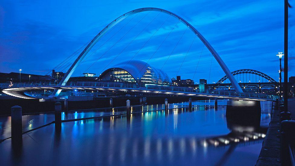 The Glasshouse in Gateshead, viewed from the east, down the River Tyne with the Gateshead Millennium Bridge in front and the Tyne Bridge in the distance behind. It's early evening or early morning and the overall tone is a crepuscular royal blue, with bridge, Glasshouse and lights all reflected in the water.