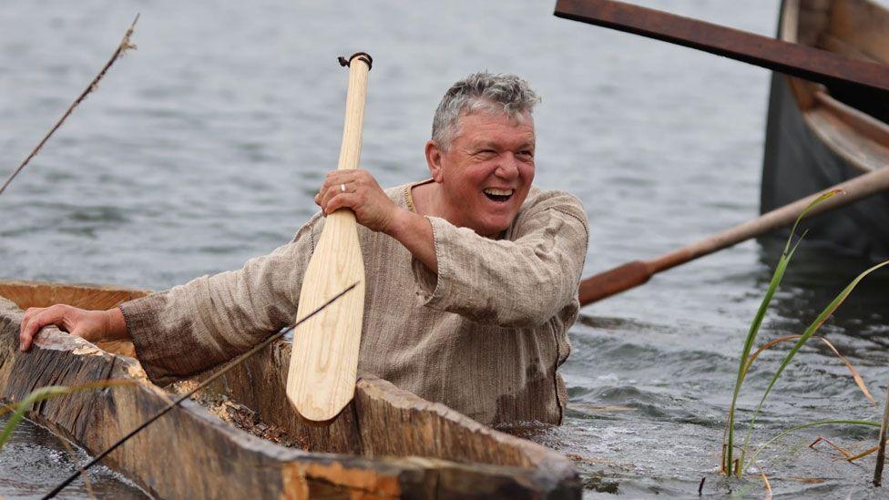 A laughing man wearing a beige tunic, waist-deep in water, towing a log boat and holding a wooden paddle