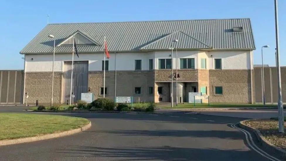 The exterior of the front entrance of Jurby Prison on a sunny day. The building is brown and cream in colour, has a grey pitched roof and flag poles flying the red Manx flag and signage in front of it. A tarmacked road leads up to it with grass on either side, and an a high perimeter fence can be seen around the complex. 