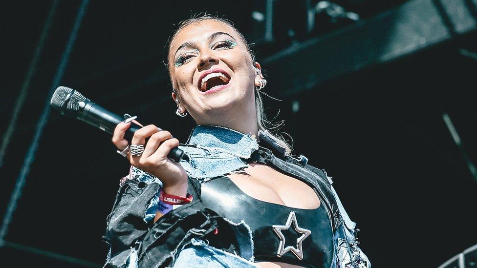 Caity Baser performing on stage. Caity is a 21-year-old woman, she has her long blonde hair tired back and wears a denim shirt over a cropped black latex top with a silver star on it. She has lots of earrings and wears pink lipstick and graphic green eyeliner and leans her head back and smiles at her audience while holding her microphone away from her with her right hand.