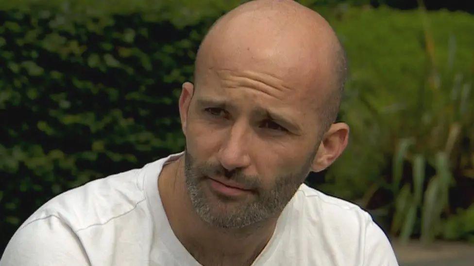 Daniel Burke wearing a white t-shirt during a TV interview. He is sat outside in a garden area.