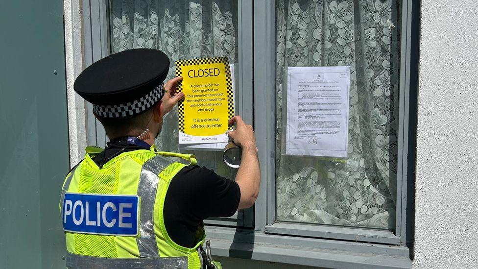 A male police officer is attaching a yellow "closed" notice to a front window of a house. The window has net curtains and another explanatory notice is attached to the other side of the window. The officer is wearing a police hat and a hi-vis yellow jacket with the word "police" across the back of it