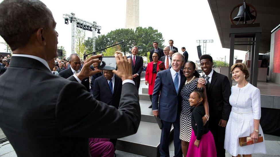 Barack Obama, left, takes a photo on a smartphone of a family with George W Bush, at a formal event.