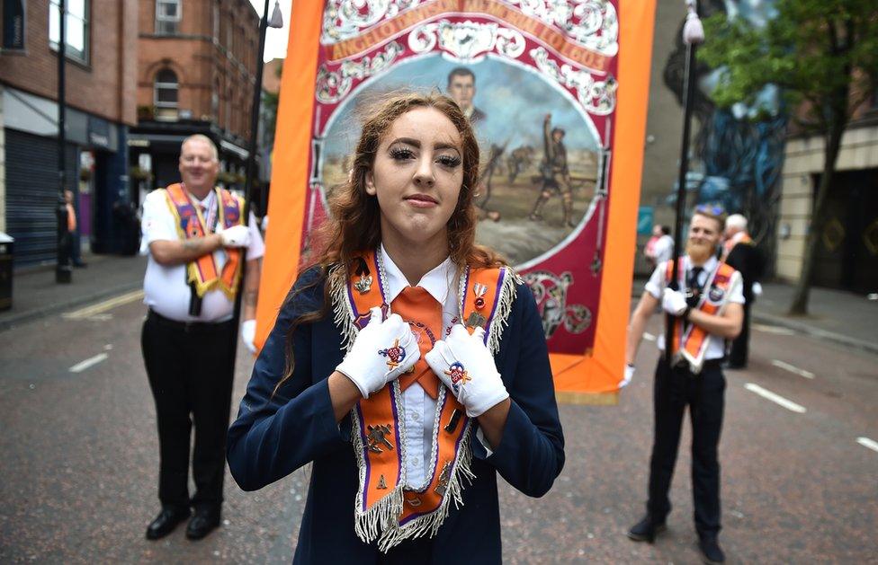 A young woman wearing an Orange Order sash with two Orangemen carrying a banner behind her