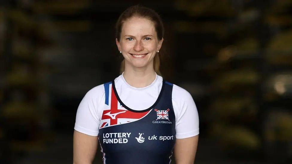 Paralympic rower Erin Kennedy poses for a photo in her Team GB rowing uniform. She is smiling, has long brown hair and small pearl earrings