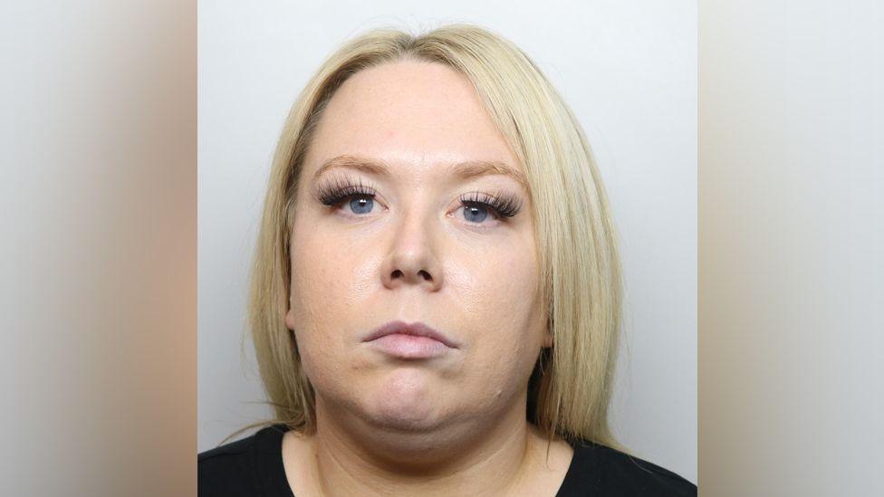 A mugshot of Chelsea Kemp. She has blonde shoulder length straight hair and blue eyes, with false eyelashes. She wears a black top and is staring at the camera.