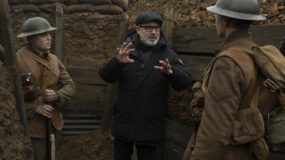 Director Sam Mendes is dressed in black, standing in a trench, and explaining something to two actors dressed as Great War soldiers