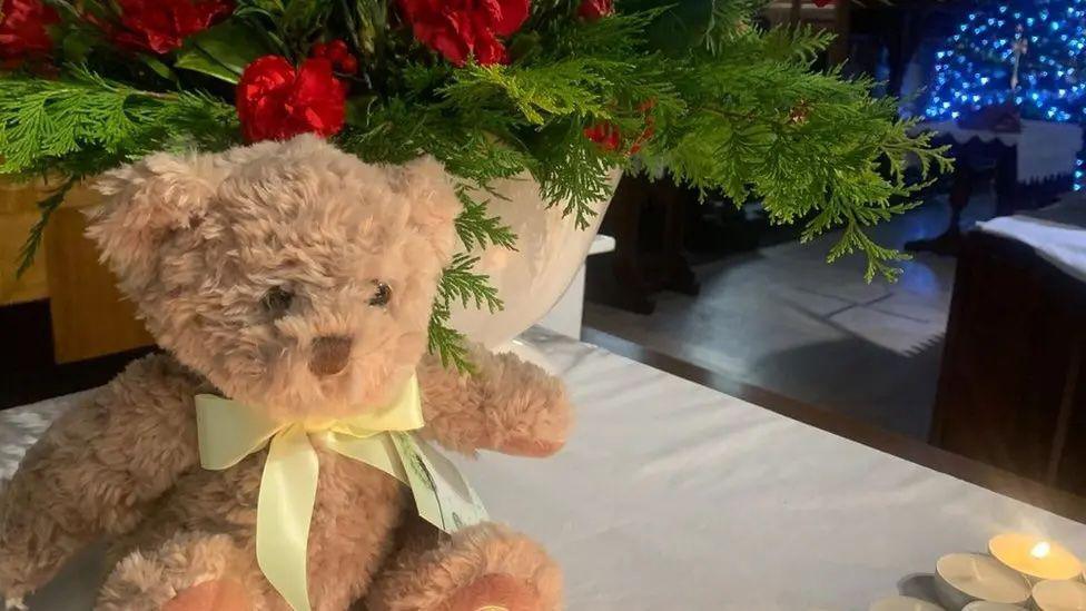 A brown teddy bear with a gold ribbon around its neck on a white tablecloth, with candles near it and a plant in the background.