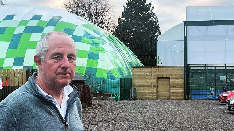 Keith Hughes has short receding grey hair. He is standing next to the green inflatable tennis courts.