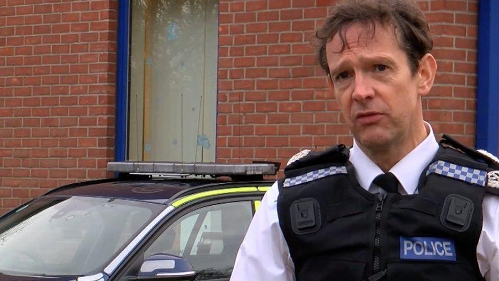 A man with dark brown hair wearing a police uniform staring just past the camera. A brick wall can be seen in the background. 