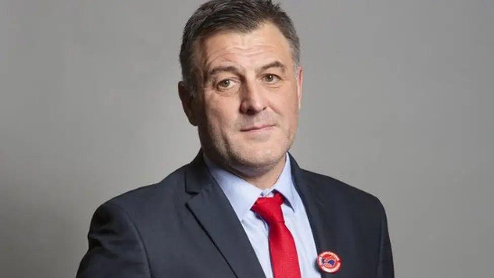 Ian Byrne has short brown hair and is wearing a navy suit with a light blue shirt and a red tie.