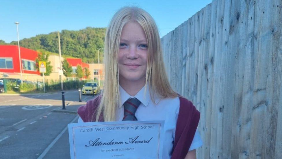 Keely Morgan yn dal tystysgrif presenoldeb ysgol ar stryd gyda'r ysgol yn y cefndir