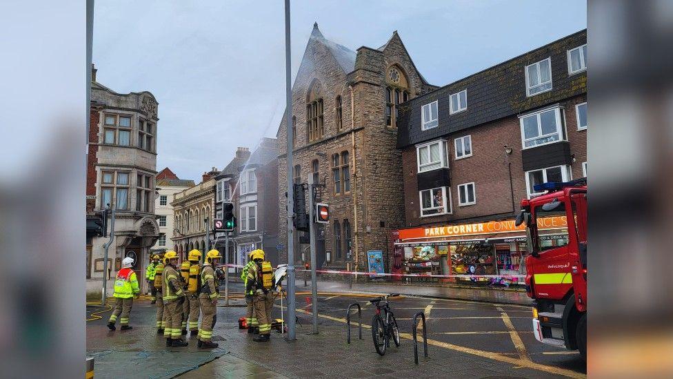 A group of firefighters standing next to a red and white cordon, with a smoking building behind it.