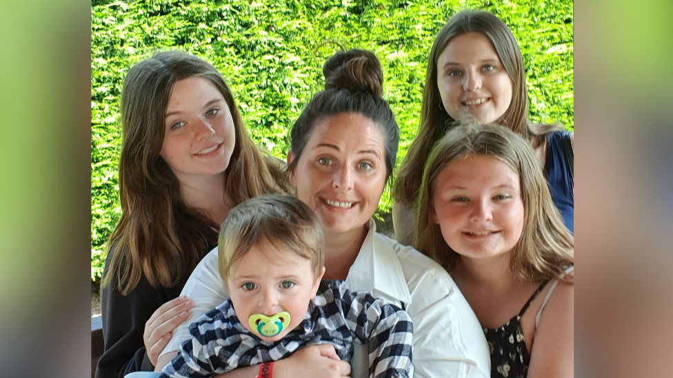 Laura-Jane Seaman has her brown her in a bun and smiles at the camera surrounded by four young children. She is wearing a white shirt and holding the youngest child.