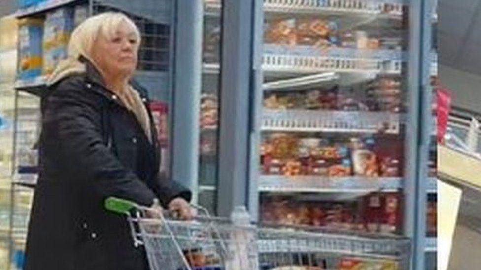 Surveillance footage of Christina Pomfrey, who has short blonde hair and is wearing a thick black coat, pushing a trolley along a supermarket aisle