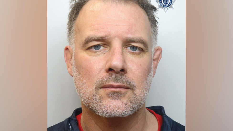 A police mugshot of Paul Whitehurst, looking at the camera. He has short, dark to white hair and a grey and white stubbled chin and cheeks. He has blue eyes. He wears a blue jacket over a red top.