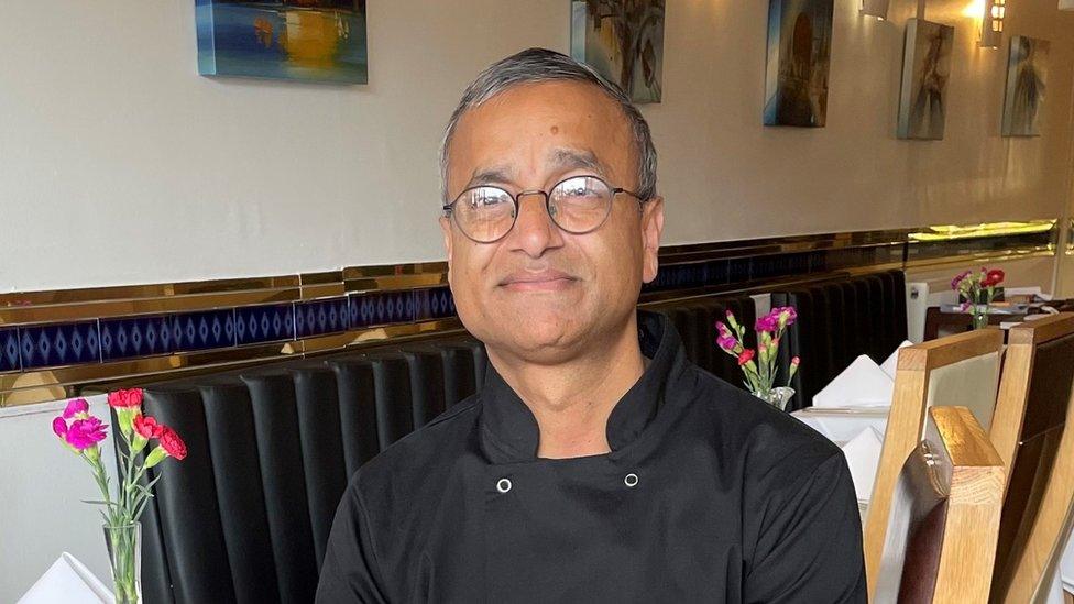 Karim Ullah is sat on a chair in his restaurant. He is an Asian man and has short dark hair that's going grey and is wearing round spectacles. He is wearing a black chef's overall and behind him you can see tables in his restaurant with purple flowers on them. There are also a few bits of artwork on the cream walls behind him.