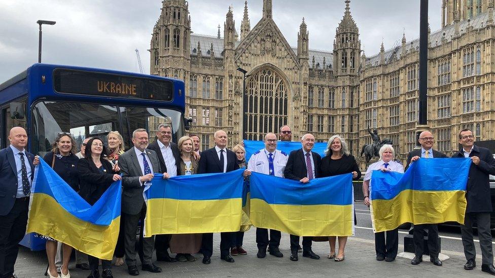 A group of people holding Ukrainian flags stand in front of a single decker bus that has been converted into a field hospital, outside the House of Lords in Westminster