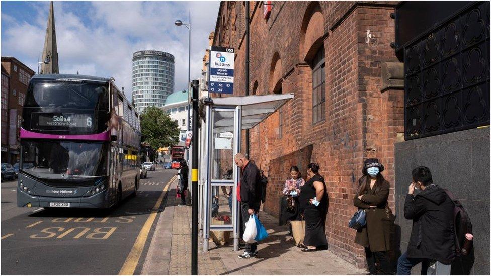 People wearing face masks waiting at a bus stop, as a bus to Solihull passes