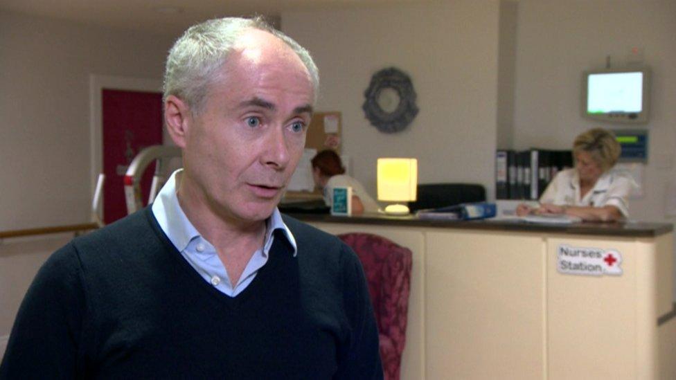 Mark King stands in the main reception of the nursing home. He has grey hair and wears a blue shirt and navy jumper.