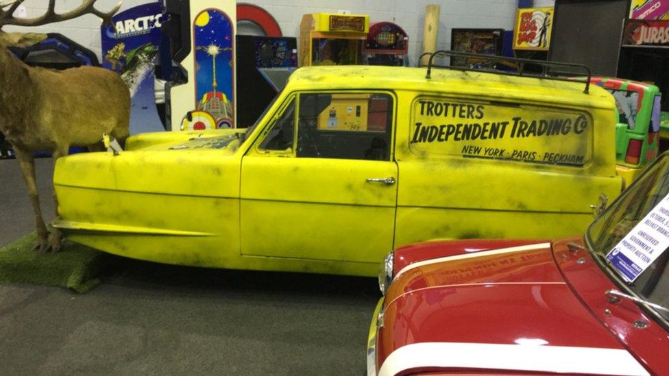 A replica Del Boy Reliant Robin car with three wheels. The yellow car has the trademark 'Trotters Independent Trading Co' logo on the side.