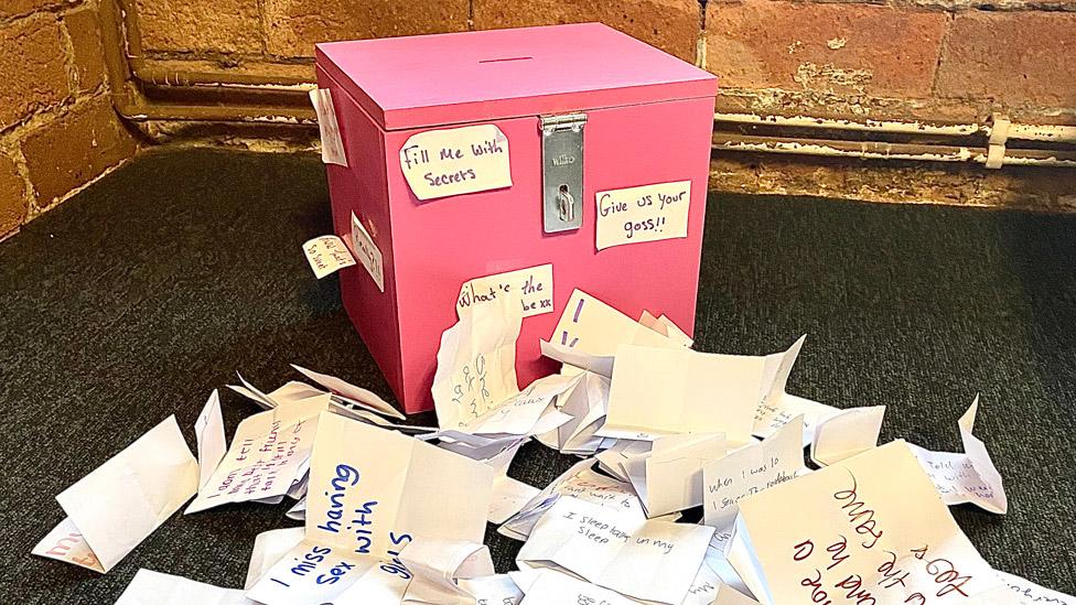 A box used to collect secrets, next to to a pile of pieces of paper with secrets written on them