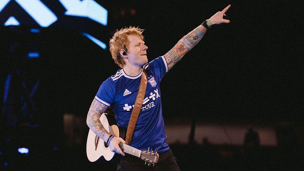 Ed Sheeran on stage wearing an Ipswich Town shirt and holding a guitar