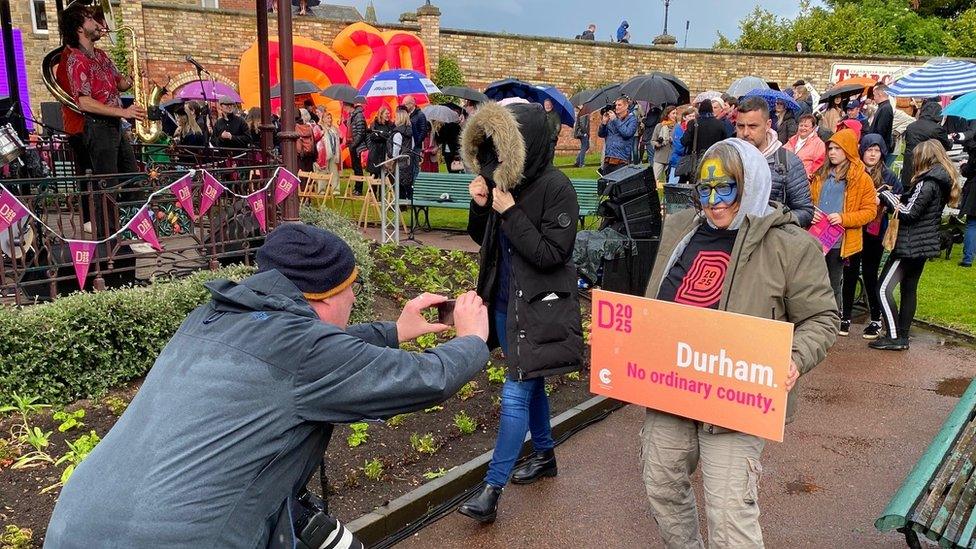 A photographer takes a picture of a woman with a blue and white yellow painted face holding a Durham 2025 banner