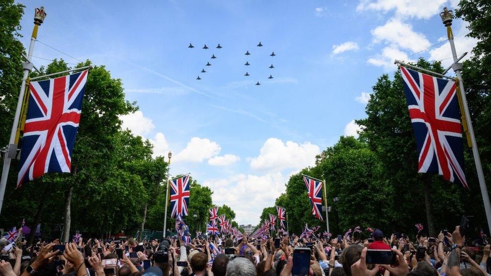 Airplanes fly in the formation of a 70 with UK flags lining the street below and crowds looking up to the sky