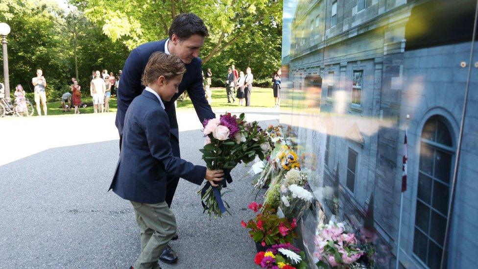 Justin Trudeau and his son leaving flowers, in smart clothing whilst people watch from behind.