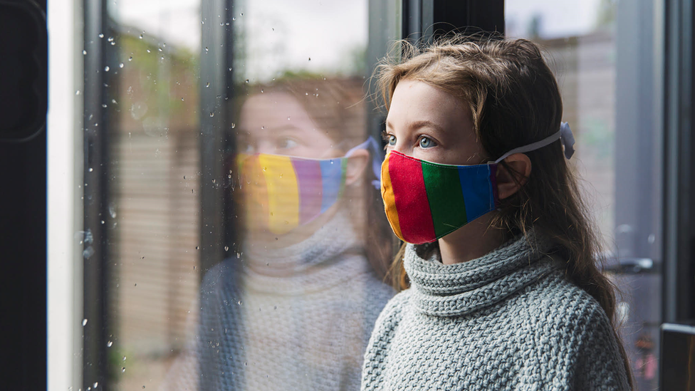 A young girl looks out the window wearing a rainbow mask.