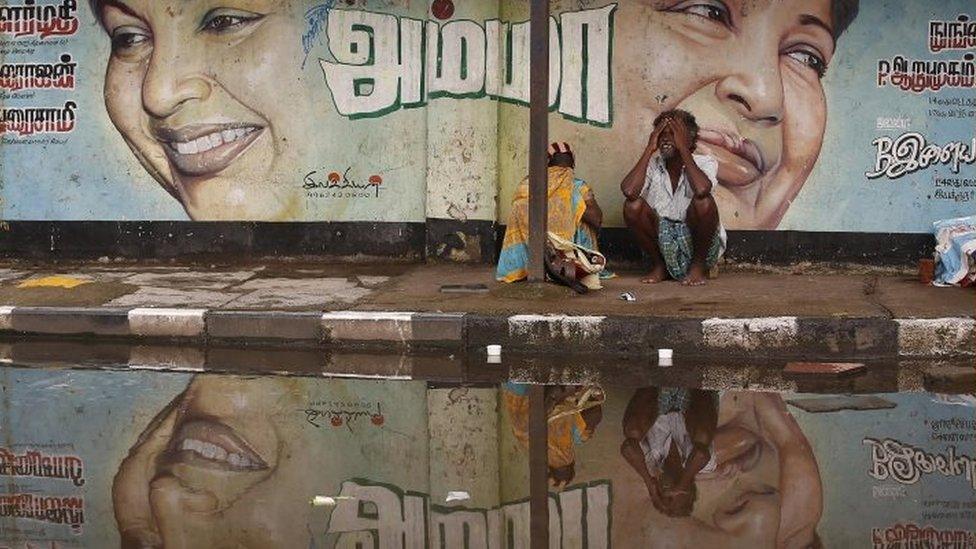 A flood-affected couple sits along a flooded roadside under a picture of Jayalalithaa Jayaram, chief minister of the southern Indian state of Tamil Nadu, in Chennai, India,