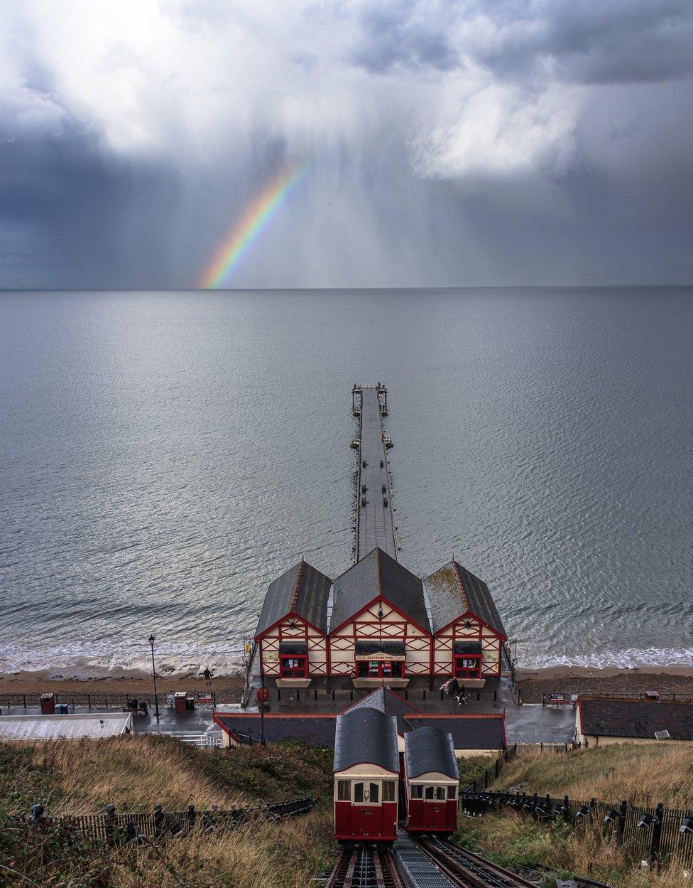 View down a vertical railway to a pier jutting out in to the sea and rainbow in the distance