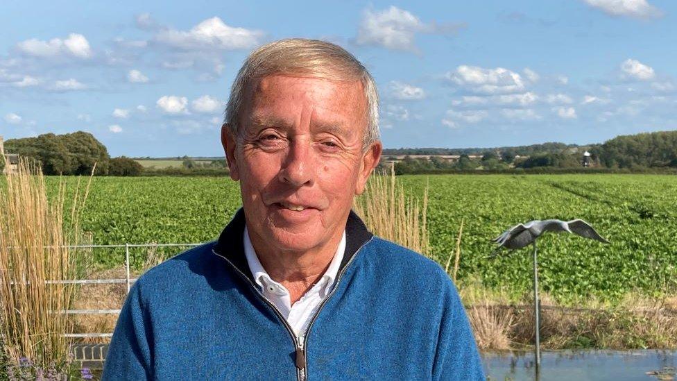 Dennis Clark, chairman of Burnham Market Parish Council. He is wearing a blue sweater and standing in front of a field.