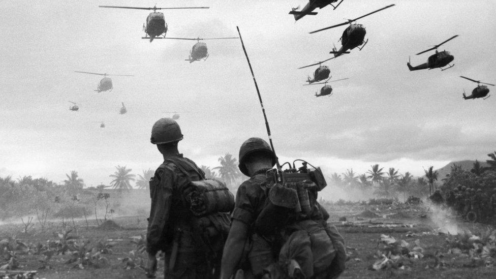 US combat helicopters fly over two American soldiers on an isolated landing zone during Operation Pershing, on the Bong Son Plain and An Lao Valley in South Vietnam, during the Vietnam War.