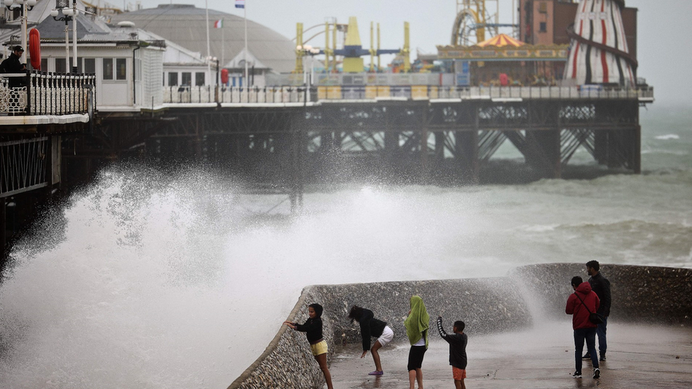 Brighton pier with large waves crashing over the sea wall. A family wearing shorts and rain jackets getting wet watching on.