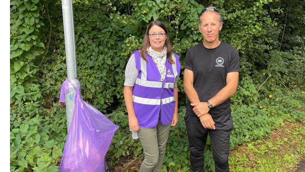 Woman in hi-viz jacket and man in black T-shirt stand next to rubbish bag