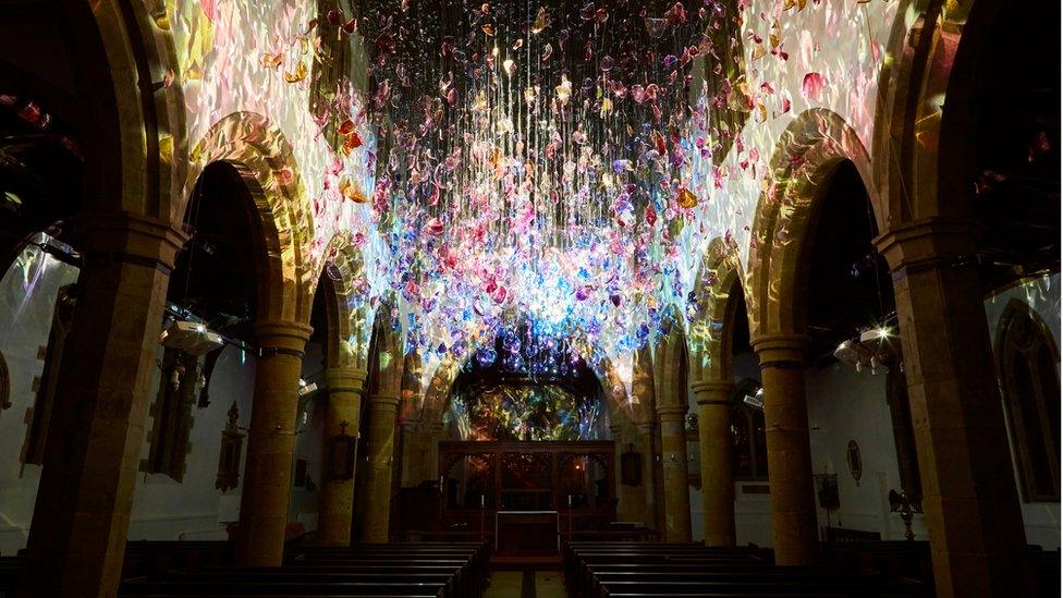 Pink, yellow, blue and purple curved glass is suspended in the air in a large church. Light shines through throwing colour all over the walls.